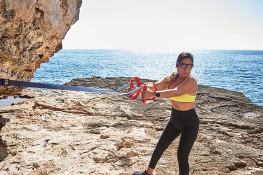FITNESS WOMAN IN SPORTS SET TRAINING WITH ELASTIC BAND, WEIGHTS, GYM EXERCISES, IN FRONT OF THE WATER.