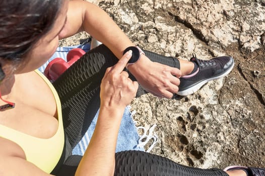 ,Latin woman, middle-aged, resting, regaining strength, eating, drinking water, after a gym session, burning calories, keeping fit, outdoors by the sea, wearing headphones and smart watch,
