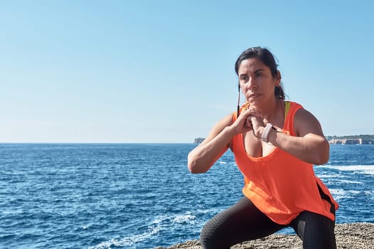Latin woman, middle-aged, wearing sportswear, training, doing physical exercises, plank, sit-ups, climber's step, burning calories, keeping fit, outdoors by the sea, wearing headphones, smart watch,