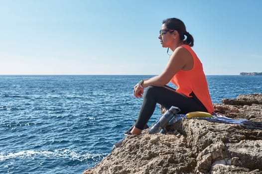 Latin woman, middle-aged, resting, regaining strength, eating, drinking water, after a gym session, burning calories, keeping fit, outdoors by the sea, wearing headphones and smart watch,