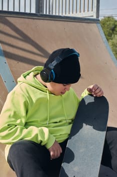 young, teenager, with a skateboard, thinking, on a rink, skateboarding, wearing headphones, green sweatshirt, black hat, swinging on a sunny day