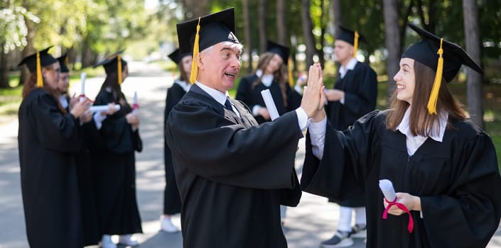 A group of graduates in robes outdoors. An elderly man and a young woman congratulate each other on their graduation