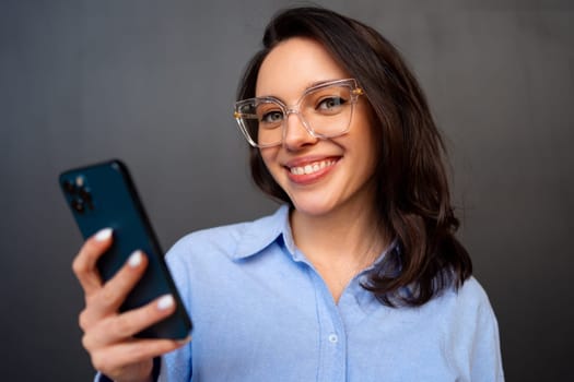 Portrait smiling woman holding smartphone in hand looking at camera, wear glasses, types text message on mobile phone, enjoys online communication, on black studio background.