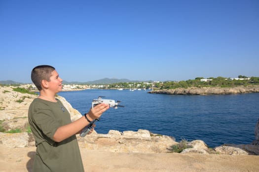 happy teenage boy, preparing and finalizing flight details for drone flight during sunny day with the mediterranean sea in the background Spain, Balearic Islands