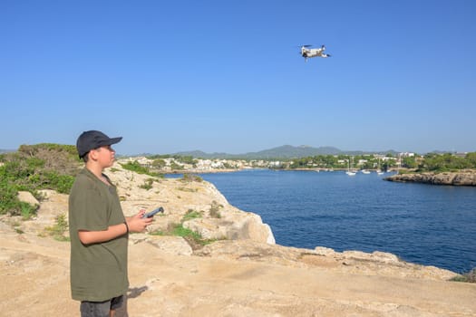 happy teen boy, flying drone on mediterranean coast, against blue sky during sunny day Spain, Balearic Islands