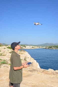 happy teen boy, flying drone on mediterranean coast, against blue sky during sunny day Spain, Balearic Islands