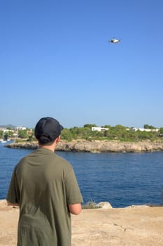 Rear view of teenage boy, flying drone on Mediterranean coast, against blue sky during sunny day Spain, Balearic Islands