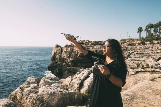 happy woman with drone, looking to the side black dress and remote control, on cliff, by the sea sunny day. technological concept,