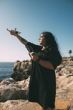 happy woman with drone, looking to the side black dress and remote control, on cliff, by the sea sunny day. technological concept,