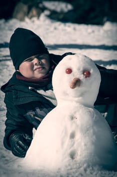 boy lying in the snow next to self-made snowman with carrot nose and tomato eyes,