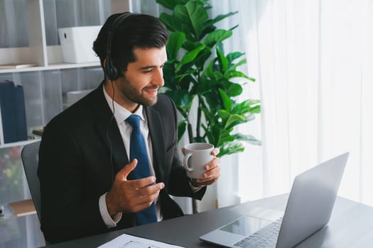 Manager of call center operator office sitting on his desk with his coffee while working on laptop. Office worker wearing headset and black suit working on customer support or telemarketing. fervent