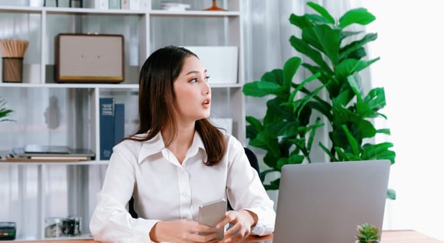 Modern and young enthusiastic businesswoman multitasks by checking her smartphone for business matters and working with laptop in her desk at the office.