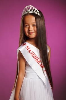 Cheerful little beauty winner posing with tiara, on purple background