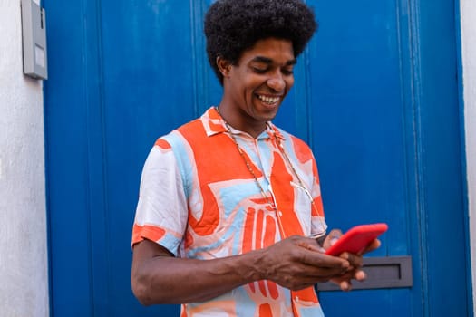 Happy black man with afro hairstyle using mobile phone to text a friend standing in the street. Blue door background. Copy space. Colourful image.