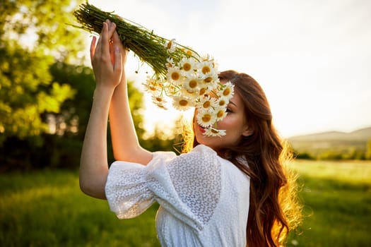 a girl in a light dress stands in nature in the sunset rays of the sun and covers her face with a bouquet of daisies. High quality photo