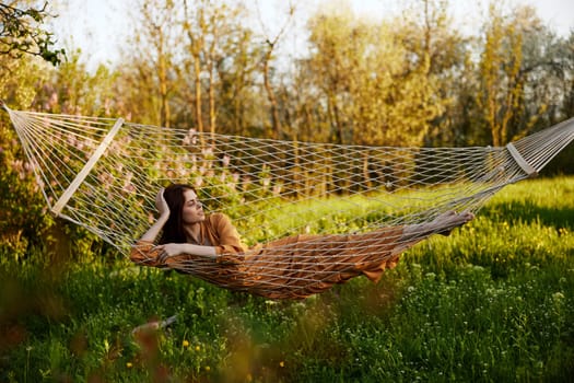 an elegant, slender woman is resting in nature lying in a mesh hammock in a long orange dress enjoying the rays of the setting sun on a warm summer day looking away. High quality photo