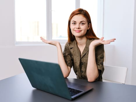 red-haired woman in overalls spreads her hands in surprise, being in the office with a laptop on the table. High quality photo