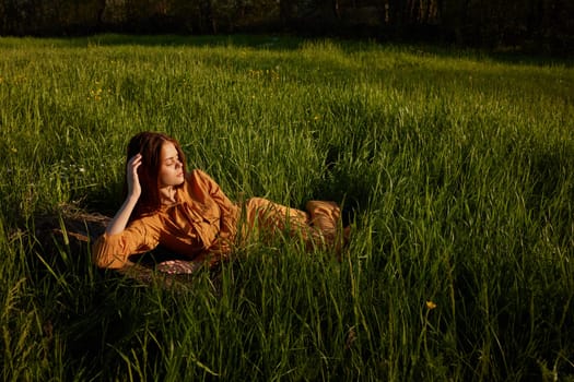 a beautiful, stylish woman lies in a long orange dress in the tall green grass illuminated by the sun rays with her eyes closed, holding her hand near her head and looking away. High quality photo
