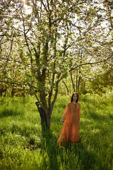 a beautiful, joyful woman stands in a long orange dress, in the countryside, near a tree blooming with white flowers, during sunset, illuminated from behind and looks into the camera. High quality photo