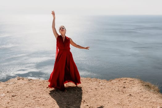 Side view a Young beautiful sensual woman in a red long dress posing on a rock high above the sea during sunrise. Girl on the nature on blue sky background. Fashion photo.