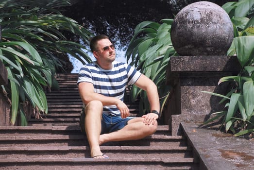 a stylish guy with glasses is sitting on the steps in a tropical garden. portrait of a young man in summer clothes. tropical country in summer. High quality photo