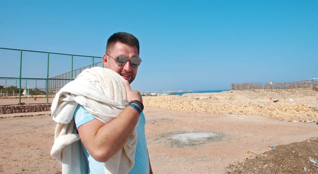 young man after a workout with a towel on his shoulder. Sports during vacation. embankment with a sea view. Sports guy goes from training. High quality photo