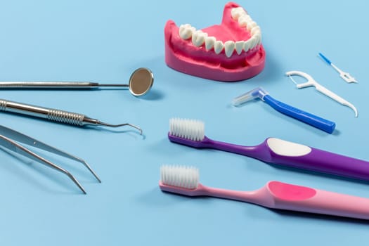 Tweezers, a mouth mirror and a dental restoration instrument with the layout of human jaw, toothbrushes, a silicone floss toothpick and interdental toothpick brushes on the blue background.
