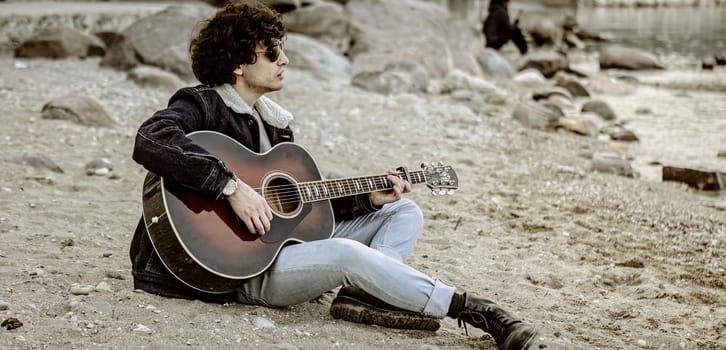 A young man sits alone on the shore, strumming his guitar and gazing out at the ocean.
