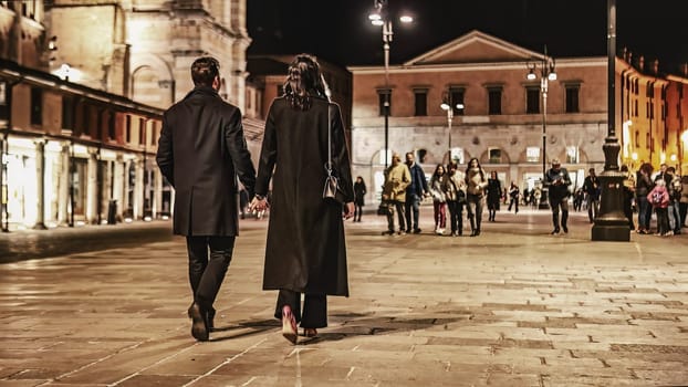 Unrecognizable couple holding hands walks on a city street at night, their silhouettes framed by city lights