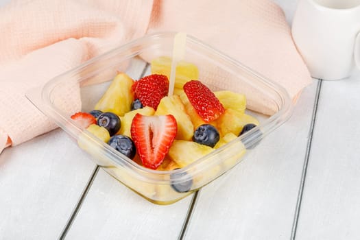 Fruit salad with pineapple, strawberries and blueberries in a plastic container on a wooden background. High quality photo