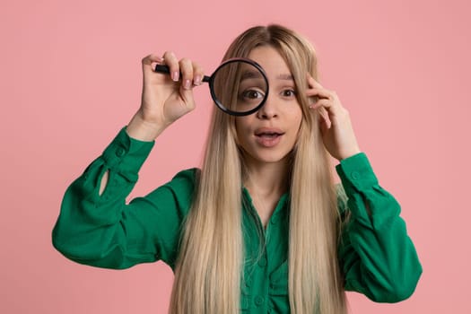 Investigator researcher scientist woman holding magnifying glass near face looking into camera with big zoomed funny eyes, searching analysing. Excited teen girl isolated on pink studio background