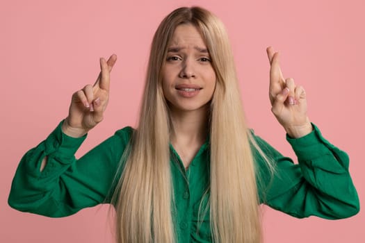 Young woman crossed fingers looking at camera asking for good luck news, wishing good education exam results dreaming about win victory in lottery jackpot. Teen girl isolated on pink studio background
