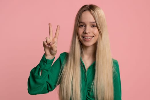 Young caucasian woman showing victory sign, hoping for success and win, doing peace gesture, smiling with kind optimistic expression. Pretty teenager girl kid isolated alone on pink studio background