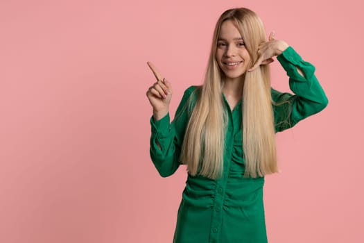 Call us, here is contact number. Pretty caucasian woman in dress looking at camera doing phone gesture like says hey you call me back conversation. Teenager girl isolated on pink background, indoor