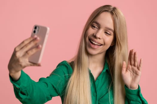 Happy Caucasian woman traveler blogger, taking selfie on smartphone, communicating video call online with family or friends, live translation stream. Pretty girl isolated on pink background, indoors