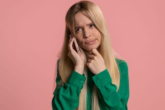 Disinterested young Caucasian woman having annoyed boring talk on smartphone. Tired sleepy adult girl talking on mobile phone with friend, making online conversation isolated on studio pink background