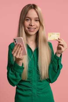 Sincere woman customer using credit bank card and smartphone while transferring money, purchases online shopping, payment. Finance and internet. Happy girl isolated alone on pink background. Vertical
