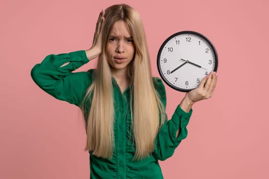 Confused woman with anxiety checking time on clock, running late to work, being in delay, deadline. Young teen girl looking at hour minutes, worrying to be punctual isolated on pink studio background