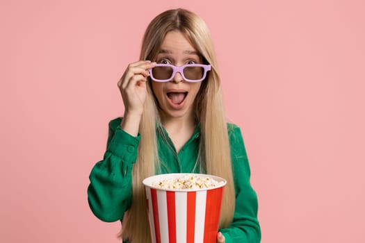 Excited pretty caucasian young woman in 3D glasses eating popcorn and watching interesting tv serial, sport game, film, online social media movie content. Teenager girl kid isolated on pink background
