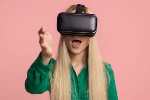 Pretty caucasian young woman using headset helmet app to play realistic simulation games. Watching virtual reality 3D 360 video. Teenager girl in VR goggles gadget isolated on pink background, indoors