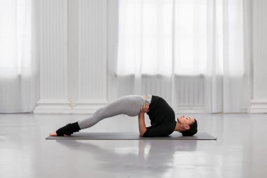 Flexible young woman doing setu bandhasana asana during yoga class in cozy bright room. The concept strengthens the neck and tones the cervical, dorsal, lumbar and sacral spine.
