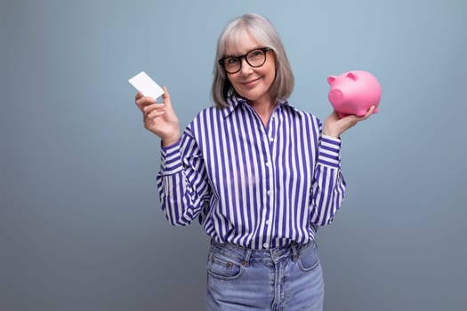 pension contribution. middle-aged woman with gray hair masters cashless payments holding a credit card and a piggy bank on a bright studio background.
