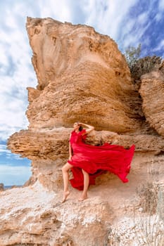 A woman in a red silk dress stands by the ocean, with mountains in the background, as her dress sways in the breeze