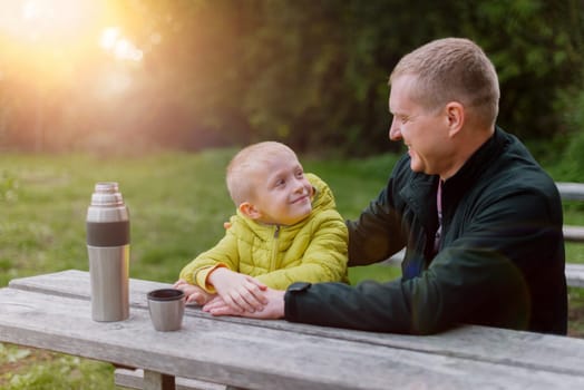 Happy family: father and child boy son playing and laughing in autumn park, sitting on wooden bench. Father and little kid having fun outdoors, playing together. Father and son sitting on a bench and talking. dad son park bench table autumn thermos.