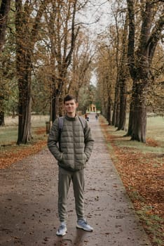 a guy in a jacket stands on an alley in the park during the fall season. Portrait oh handsome teen guy, young man in hoodie, down jacket standing, walking in beautiful golden autumn park, looking at camera. Natural background, colourful leaves, trees. Portrait of young smiling man in casual jacket looks at camera on autumn nature background in countryside or in park. Concept of style, walking in fresh air and unity with nature