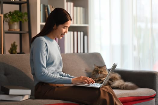 Nice asian woman using laptop computer on couch and smiling moment with domestic cat. High quality photo