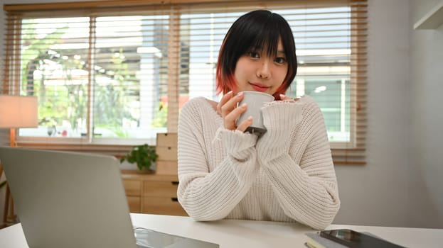 Pleasant young asian woman drinking coffee, sitting in front of laptop computer at home office desk.