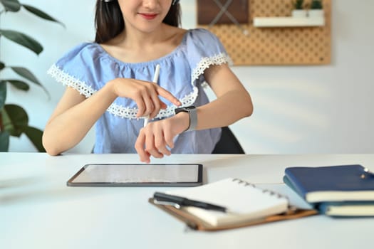 Cropped shot of young woman checking time on her smartwatch and using digital tablet at working desk.