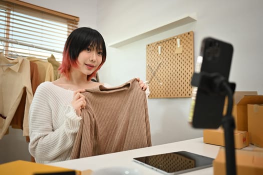 Young woman online seller showing clothes in front of smartphone camera, selling clothes online by streaming.