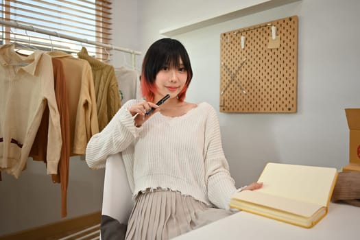 Young female fashion designer standing near clothing rack, working on new womenswear collection in modern studio.